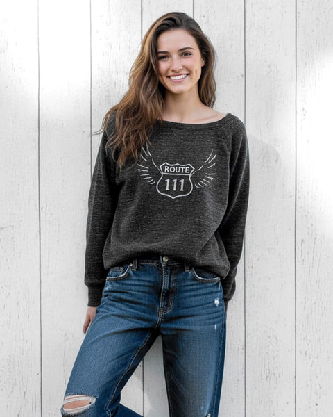Woman wearing a heather charcoal wide‑neck sweatshirt with a vintage 111 angel number design featuring wings.