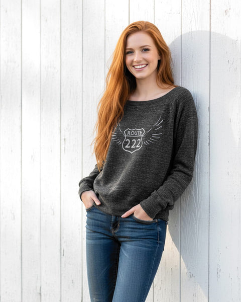 Woman wearing a heather charcoal wide‑neck sweatshirt with a vintage 222 angel number design featuring wings.